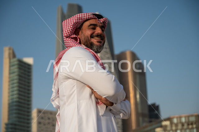 النظر الى الكاميرا بإيماءات السرور والسعادة، صورة من الجانب لرجل عربي خليجي سعودي يرتدي الشماغ والثوب التقليدي ويقف بايدي مكتوفة، الفن الهندسي المعماري للأبراج وناطحات السحاب في الرياض، النمو والتطور العمراني في السعودية، خلفية مركز الملك عبد الله المالي