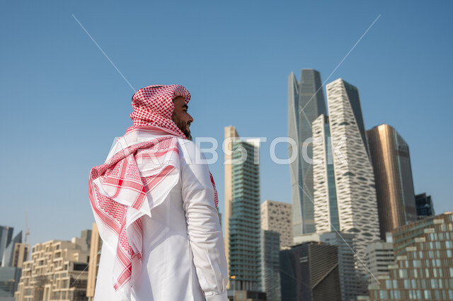 الفن الهندسي المعماري لمركز الملك عبد الله المالي (كافد)، صورة من الخلف لرجل عربي خليجي سعودي يرتدي الثوب التقليدي والشماغ وينظر الى المباني المالية والمنشآت الإقتصادية، النمور والتطور العمراني في مدينة الرياض، الأبراج وناطحات السحاب في السعودية