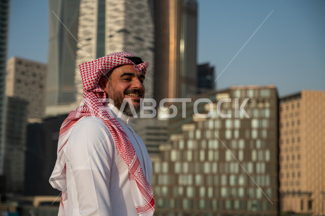 النمو والتطور العمراني في السعودية، المباني المالية والمنشآت الإقتصادية، صورة من الجانب لرجل عربي خليجي سعودي يرتدي الزي التقليدي وينظر الى الكاميرا بسعادة، الفن الهندسي المعماري للأبراج وناطحات السحاب في الرياض، خلفية مركز الملك عبد الله المالي (كافد) 