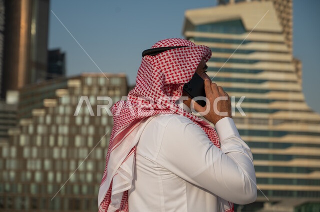 التواصل والتحدث مع الأشخاص من خلال الجوال، صورة من الجانب لرجل عربي خليجي سعودي يرتدي الزي التقليدي ويجري مكالمة من خلال الهاتف المحمول، الفن الهندسي المعماري للأبراج وناطحات السحاب في الرياض، النمو والتطور العمراني للمباني المالية والمنشآت الإقتصادية