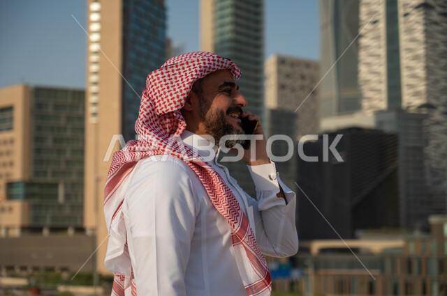الفن الهندسي المعماري للأبراج وناطحات السحاب في الرياض، صورة من الجانب لرجل عربي خليجي سعودي مبتسم يرتدي الزي التقليدي ويجري مكالمة من خلال الهاتف المحمول، النمو والتطور العمراني للمباني المالية والمنشآت الإقتصادية، التواصل والتحدث مع الأشخاص من خلال الجوال