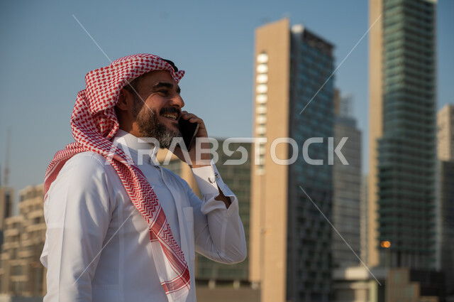النمو والتطور العمراني للمباني المالية والمنشآت الإقتصادية، صورة من الجانب لرجل عربي خليجي سعودي مبتسم يرتدي الزي التقليدي ويجري مكالمة من خلال الهاتف المحمول، الفن الهندسي المعماري للأبراج وناطحات السحاب في الرياض، التواصل والتحدث مع الأشخاص من خلال الجوال