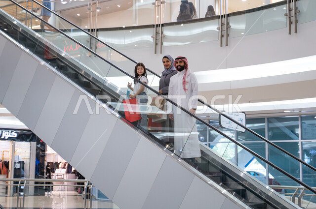مفهوم الأسره السعيده, الصعود والانتقال إلى الطابق الآخر باستخدام السلم الكهربائي, عائلة عربيه خليجيه سعوديه يحملون أكياس التسوق الملونه, التجول والتسوق في إحدى المولات التجاريه في المملكه العربيه السعوديه, الوقوف والنظر إلى الكاميرا بإيماءات السرور والسعاده