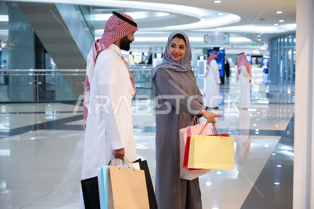 أهميه الاستفاده من العروض والتخفيضات الموسميه, مشاركة الزوجين في اختيارات التسوق وتبادل الآراء, رجل وإمرأه عربيان سعوديان خليجيان يحملان أكياس التسوق الملونه, ارتداء الزي السعودي التقليدي, الاستمتاع في قضاء وقت ممتع في إحدى المولات التجاريه في المملكه