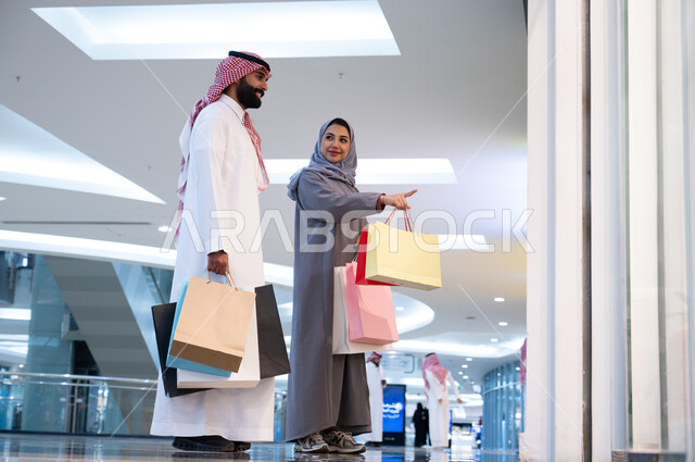 مشاركة الزوجين في اختيارات التسوق وتبادل الآراء, رجل وإمرأه عربيان سعوديان خليجيان يحملان أكياس التسوق الملونه, ارتداء الزي السعودي التقليدي, الاستمتاع في قضاء وقت ممتع في إحدى المولات التجاريه في المملكه, الاستفاده من العروض والتخفيضات الموسميه