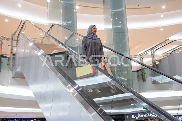 استخدام الدرجات والسلالم الكهربائية في المولات التجاريه, إمرأة عربيه خليجيه سعوديه مبتسمه محجبه ترتدي العباءة تحمل الأكياس الملونه, التجول في الأسواق السعوديه, الاستمتاع بقضاء وقت ممتع في التسوق والشراء