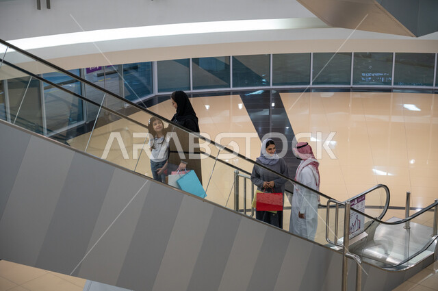 سهولة الانتقال من طابق لآخر باستخدام السلالم الكهربائية, صورة من الأعلى لمجموعه من المواطنين العرب الخليجيين السعوديين يقومون باستخدام الدرج الكهربائي, متعه التسوق في المولات التجاريه واختيار المستلزمات والهدايا, الاستفاده من العروض والتخفيضات الموسميه