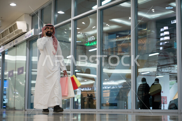 استخدام الهاتف المحمول في أسواق السعوديه, رجل عربي خليجي سعودي يرتدي الثوب التقليدي والشماغ يحمل أكياس التسوق الملونه ويقوم بإجراء مكالمة هاتفيه عبر الجوال, التجول في إحدى المولات التجاريه في المملكه, الاستمتاع في التسوق وشراء المستلزمات والهدايا