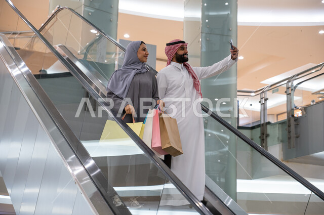 Choosing and purchasing supplies and gifts, the importance of spending quality time with the family, a Saudi-Gulf Arab couple walking around in one of the malls, going down the electronic escalator and taking souvenir selfies via mobile phone, the fun of shopping in the Saudi markets.