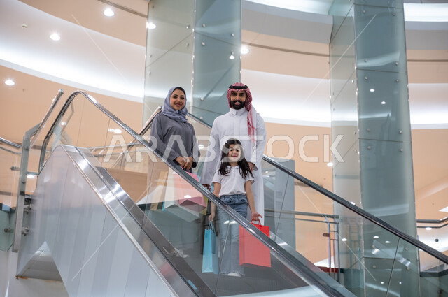 مفهوم الأسره السعيده, النزول والانتقال إلى الطابق الآخر باستخدام السلم الكهربائي, عائلة عربيه خليجيه سعوديه يحملون أكياس التسوق الملونه, التجول والتسوق في إحدى المولات التجاريه في المملكه العربيه السعوديه, الوقوف والنظر إلى الكاميرا بإيماءات السرور والسعاده