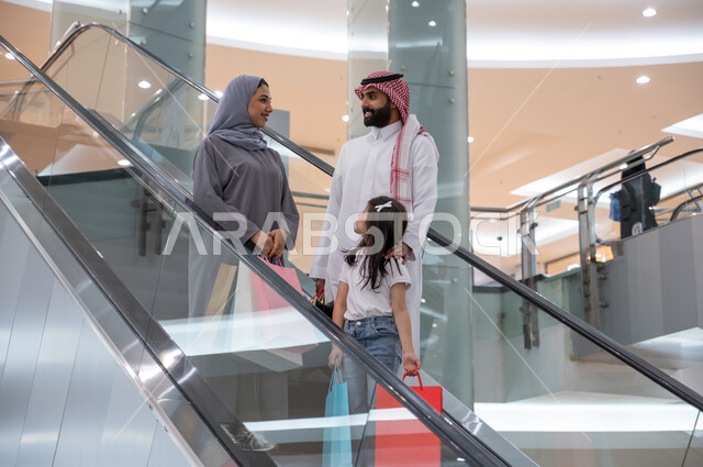 النزول والانتقال باستخدام السلم الكهربائي, عائلة عربية خليجية سعودية سعيدة تحمل أكياس التسوق و تتجول في مركز التسوق، الاستمتاع بالشراء وانتقاء المستلزمات والهدايا، الاستفادة من العروض والخصومات، قضاء وقت ممتع في التسوق من المول تجاري