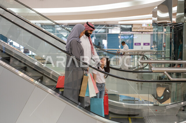 Spending an enjoyable time shopping in the mall, going up and down using the escalator, a side photo of a happy Saudi Arabian Gulf family carrying shopping bags and walking around the mall, enjoying buying and selecting supplies and gifts, taking advantage of offers and discounts.