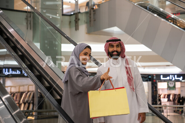 النظر والإشارة إلى شيئ ما بإيماءات السرور, مشاركة الزوجين في اختيارات التسوق وتبادل الآراء, رجل وإمرأه عربيان سعوديان خليجيان يستمتعان بالتسوق في إحدى المولات التجاريه في المملكة, استخدام السلالم الكهربائية للانتقال للطابق الآخر