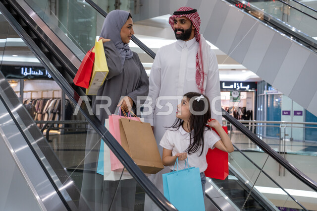 الانتقال والنزول باستخدام السلم الكهربائي, عائلة عربية خليجية سعودية سعيدة تحمل أكياس التسوق و تتجول في مركز التسوق، الاستمتاع بالشراء وانتقاء المستلزمات والهدايا، الاستفادة من العروض والخصومات، قضاء وقت ممتع في التسوق من المول تجاري