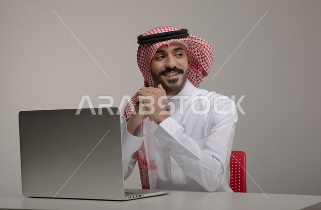 Using modern technologies in remote work, professionalizing computer and information technology jobs, portrait of a Saudi Gulf Arab man wearing traditional clothing, smiling and using a laptop, completing office tasks through the laptop, gray background