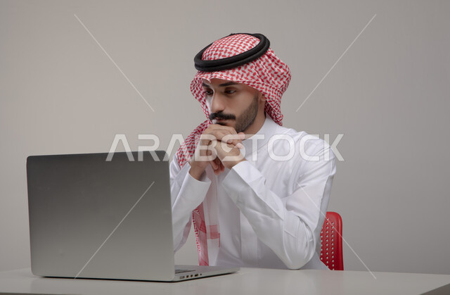 إحتراف وظائف الحاسوب وتكنولوجيا المعلومات، بورتريه لرجل عربي خليجي  سعودي يرتدي الزي التقليدي ويستخدم جهاز الحاسوب المحمول، تصفح الإنترنت والعمل عن بعد، إنجاز المهام المكتبية من خلال اللابتوب، إيماءات وجه ويدين تدل على التفكير والتحليل ، خلفية رمادية