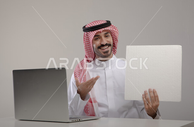 الإشارة إلى الورقة بإيماءات وجه ويدين تدل على النجاح والفرح , احتراف وظائف الحاسوب وتكنولوجيا المعلومات، بورتريه لرجل عربي خليجي سعودي يرتدي الزي التقليدي ويحمل بيده ورقة بيضاء فارغة ، تصفح الإنترنت والعمل عن بعد، إنجاز المهام المكتبية من خلال اللابتوب،  خلفية رمادية
