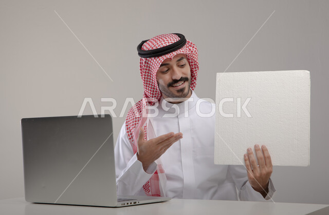 استخدام الحاسوب المحمول لإنجاز المهام المكتبية ، بورتريه لرجل عربي خليجي  سعودي يرتدي الزي التقليدي ويحمل بيده ورقة بيضاء فارغة ينظر إليها ، تصفح الإنترنت والعمل عن بعد، إيماءات وجه تدل على النجاح والسعادة ,  خلفية رمادية