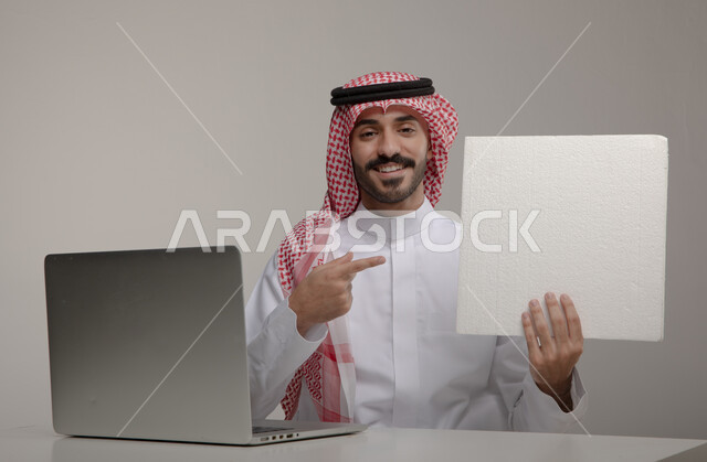 التسويق الالكتروني باستخدام اللابتوب ، إنجاز المهام المكتبية , بورتريه لرجل عربي خليجي سعودي يرتدي الزي التقليدي ويحمل بيده ورقة بيضاء فارغة ، تصفح الإنترنت والعمل عن بعد، إيماءات وجه ويدين تدل على الإشارة إلى الورقة بسعادة ، خلفية رمادية