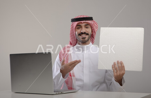 Facial and hand gestures indicating pointing to the paper, professional computer and information technology jobs, portrait of a Saudi Gulf Arab man wearing traditional clothing and holding a blank white sheet of paper in his hand, browsing the Internet and working remotely, completing office tasks through the laptop, gray background