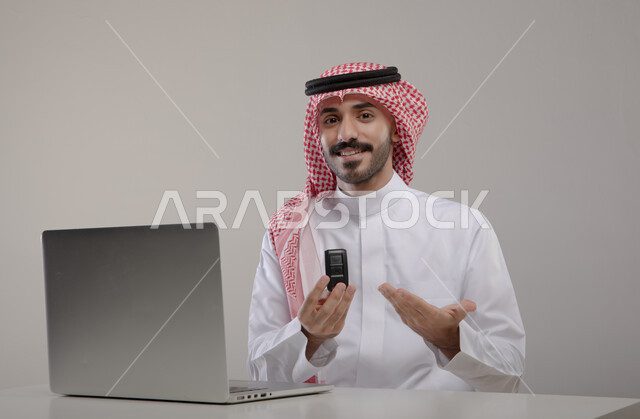تكنولوجيا وتقنيات حديثة ، بورتريه لرجل عربي خليجي سعودي يرتدي الزي التقليدي ويحمل بيده مفتاح سيارة ،إيماءات وجه ويدين مختلفة ,  تصفح الإنترنت والعمل عن بعد، إنجاز المهام المكتبية من خلال اللابتوب، استخدام جهاز الحاسوب المحمول ، خلفية رمادية
