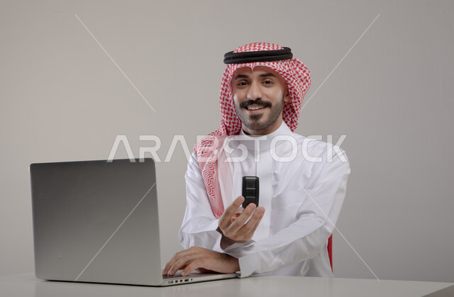 Completing office tasks through the laptop, mastering computer and information technology jobs, a portrait of a smiling Saudi Arabian Gulf man wearing traditional clothing and holding a car key in his hand, browsing the Internet and working remotely, pressing keyboard buttons, gray background