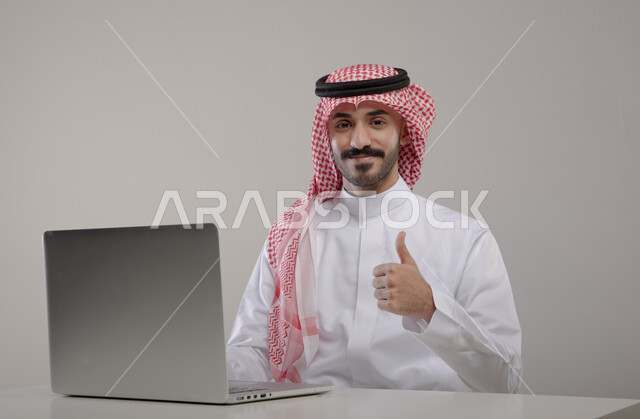  تصفح الإنترنت والعمل عن بعد عبر اللابتوب ، إحتراف وظائف الحاسوب وتكنولوجيا المعلومات، بورتريه لرجل عربي خليجي سعودي يرتدي الزي التقليدي و يرفع أصبعه الإبهام بإيماءات و تعابير وجه  تدل على الجودة و الإعجاب ، الضغط على أزرار لوحة المفاتيح، خلفية رمادية