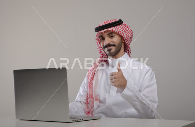 إحتراف وظائف الحاسوب وتكنولوجيا المعلومات، بورتريه لرجل عربي خليجي سعودي يرتدي الزي التقليدي و يرفع أصبعه الإبهام بإيماءات و تعابير وجه  تدل على الجودة و الإعجاب ، تصفح الإنترنت والعمل عن بعد عبر اللابتوب   ، الضغط على أزرار لوحة المفاتيح، خلفية رمادية