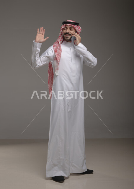 الترحيب وإيماءات تدل على السلام , استخدام الهاتف المحمول في الاتصال والتواصل مع الآخرين ، دردشة ومحادثات وتعارف، بورتريه لرجل عربي خليجي سعودي يرتدي الزي التقليدي ويجري مكالمة هاتفية من خلال الجوال، صوة الجسم بالطول الكامل , خلفية رمادية .