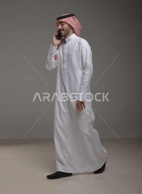 مكالمة خاصة بالعمل , استخدام الهاتف المحمول في الاتصال والتواصل مع الآخرين ، دردشة ومحادثات وتعارف، بورتريه لرجل عربي خليجي سعودي يرتدي الزي التقليدي ويجري مكالمة هاتفية من خلال الجوال، البحث عن عمل عبر الانترنت , صورة الجسم بالطول الكامل , خلفية رمادية .