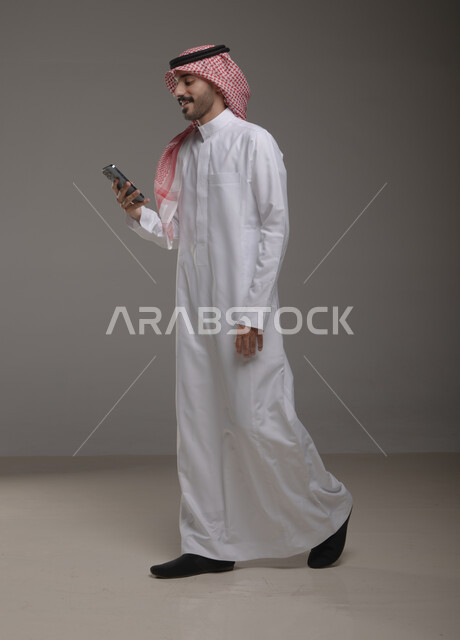 التسويق الالكتروني باستخدام الهاتف المحمول , استخدام التقنيات الحديثة في العمل ، بورتريه لرجل عربي خليجي سعودي يرتدي الزي التقليدي ويمسك بيده الجوال ، إيماءات تدل على السير , الدردشة والتواصل مع الآخرين , صورة الجسم بالطول الكامل , خلفية رمادية .