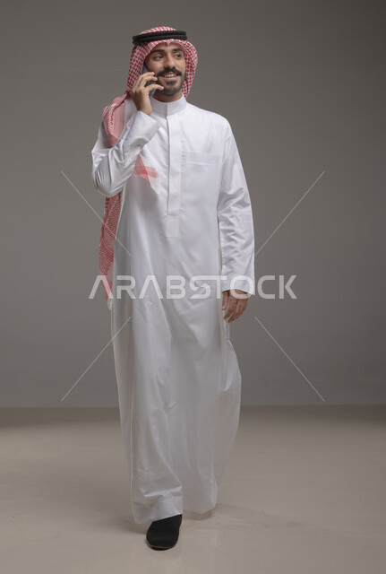 تواصل اجتماعي واتصالات عبر الهاتف المحمول , دردشات ومحادثات وتعارف، بورتريه لرجل عربي خليجي سعودي يرتدي الزي التقليدي ويجري مكالمة هاتفية من خلال الجوال، إيماءات وجه تدل على السعادة , صورة الجسم بالطول الكامل , خلفية رمادية .