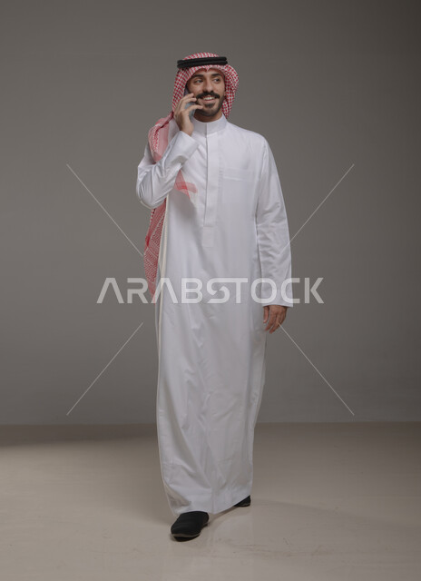 Communications and phone calls via mobile phone, chatting and communicating with family and friends, portrait of a Saudi Gulf Arab man wearing traditional clothing and using a mobile phone, gestures indicating walking and happiness, full-length body photo, gray background