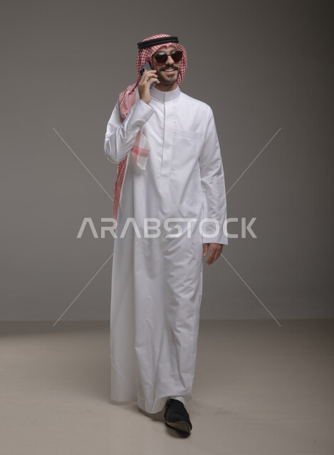 مكالمات هاتفية باستخدام الهاتف المحمول ,  التواصل مع الأهل والأصدقاء ،  بورتريه لرجل عربي خليجي سعودي يرتدي الزي التقليدي والنضارة الشمسية ويبتسم ، إيماءات تدل على السير , صورة الجسم بالطول الكامل , خلفية رمادية .