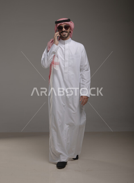 دردشة ومحادثات والتواصل مع الآخرين ،استخدام الهاتف المحمول في الاتصال , بورتريه لرجل عربي خليجي سعودي يرتدي الزي التقليدي والنضارة الشمسية وينظر إلى الكاميرا ,  إجراء مكالمة هاتفية من خلال الجوال، صورة الجسم بالطول الكامل , خلفية رمادية .
