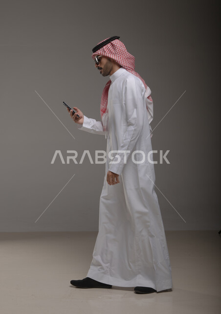 استخدام الهاتف المحمول في التواصل مع الأفراد، دردشة ومحادثات وتعارف، بورتريه لرجل عربي خليجي سعودي يرتدي الزي التقليدي ويستخدم الجوال، تصفح الانترنت والتسوق الالكتروني , صورة الجسم بالطول الكامل ,  خلفية رمادية .