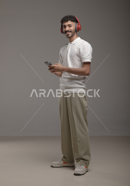 Using modern headphone technology, listening to favorite music clips, a portrait of a handsome Saudi Gulf Arab young man wearing a casual outfit, holding a mobile phone in his hand and wearing an audio headset, looking at the camera with pleased gestures, full-length body portrait, gray background