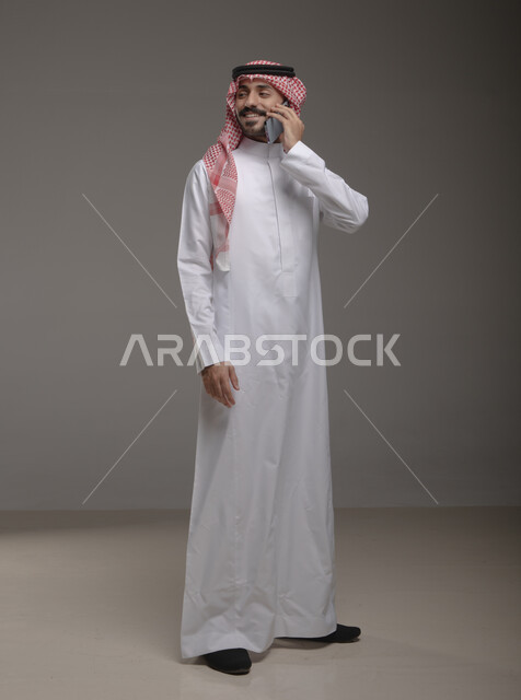 التحدث مع الأهل والأصدقاء ، دردشات ومحادثات وتعارف، بورتريه لرجل عربي خليجي سعودي يرتدي الزي التقليدي ويجري مكالمة هاتفية من خلال الجوال، إيماءات وجه تدل على السعادة , صورة الجسم بالطول الكامل , خلفية رمادية .