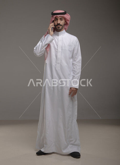 الوقوف باستقامة والنظر إلى الكاميرا , استخدام الهاتف المحمول في الاتصال والتواصل مع الأفراد، دردشة ومحادثات وتعارف، بورتريه لرجل عربي خليجي سعودي يرتدي الزي التقليدي ويجري مكالمة هاتفية من خلال الجوال، صورة الجسم بالطول الكامل , خلفية رمادية .