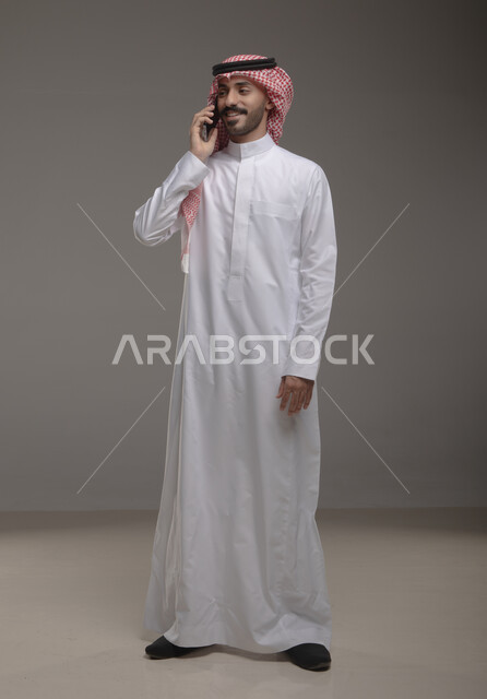 البحث عن عمل عبر وسائل التواصل الاجتماعي , استخدام الهاتف المحمول في الاتصال والتواصل مع الأفراد، دردشة ومحادثات وتعارف، بورتريه لرجل عربي خليجي سعودي يرتدي الزي التقليدي ويجري مكالمة هاتفية من خلال الجوال، صورة الجسم بالطول الكامل ,  خلفية رمادية .