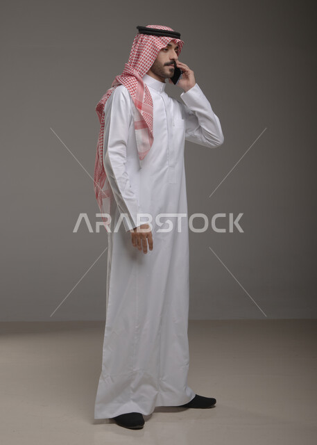 تقنيات وأجهزة حديثة , استخدام الهاتف المحمول في الاتصال والتواصل مع الأفراد، دردشة ومحادثات وتعارف، بورتريه لرجل عربي خليجي سعودي يرتدي الزي التقليدي ويجري مكالمة هاتفية من خلال الجوال،صورة الجسم بالطول الكامل ,  خلفية رمادية .