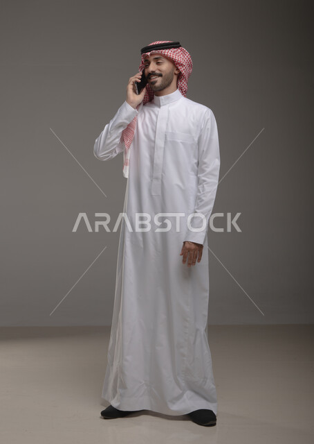 محادثات وترتيبات عمل عبر الهاتف المحمول ,التواصل مع الأصدقاء والأهل ، بورتريه لرجل عربي خليجي سعودي يرتدي الزي التقليدي ويجري مكالمة هاتفية من خلال الجوال،صورة الجسم بالطول الكامل , خلفية رمادية .
