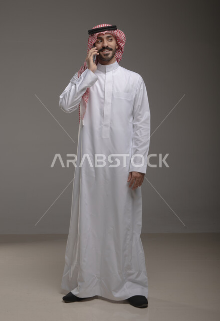 Online shopping, spending time communicating through social media applications, portrait of a Saudi Gulf Arab man wearing traditional clothing and making a phone call through a mobile phone, full-length body photo, gray background