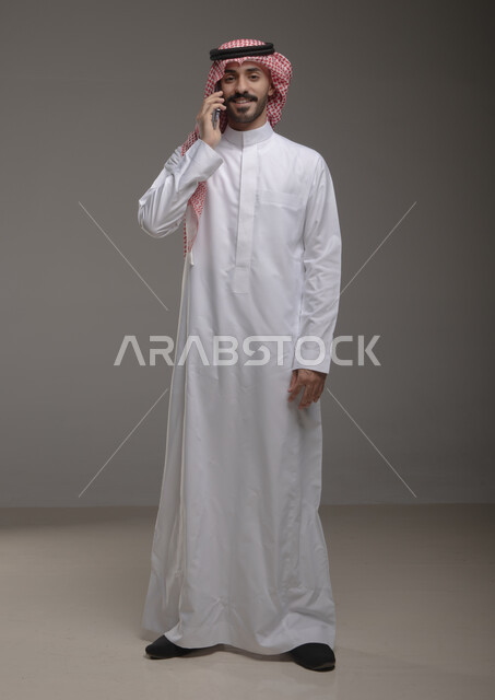 الأناقة والاهتمام بالمظهر الخارجي , استخدام الهاتف المحمول في الاتصال والتواصل مع الأفراد، البحث عن عمل عبر الانترنت ، بورتريه لرجل عربي خليجي سعودي يرتدي الزي التقليدي ويجري مكالمة هاتفية من خلال الجوال، صورة الجسم بالطول الكامل , خلفية رمادية .