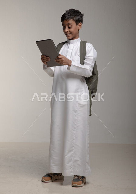 دمج التقنية والتكنولوجيا مع التعلم، البحث عن معلومات مفيدة وهامة، بورتريه لفتى عربي خليجي سعودي مبتسم يرتدي الثوب التقليدي وحقيبة الظهر يحمل بيده جهازالتابلت، متابعة مقاطع ثقافية وحضور دورات تعليمية الكترونية، صورة الجسم بالطول الكامل، خلفية رمادية