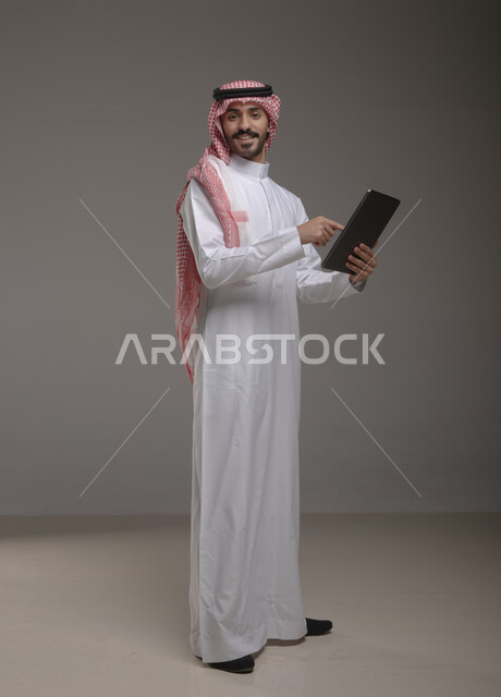 الضغط والنقر باستخدام حاسة اللمس , بورتريه لرجل عربي خليجي سعودي يرتدي الزي  التقليدي يحمل بيده  التابليت و ينظر إلى الكاميرا , التسويق الرقمي و ترويجات إعلانية  ، تطبيقات الأجهزة اللوحية  ، صورة الجسم بالطول الكامل , خلفية رمادية .
