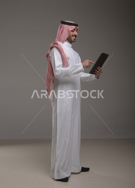 استخدام الأجهزة التقنية الحديثة , بورتريه لرجل عربي خليجي سعودي يرتدي الزي  التقليدي يحمل بيده  التابليت  ،الدردشة مع الأهل والأصدقاء  , تصفح برامج التواصل الاجتماعي   ، تطبيقات الأجهزة اللوحية  ، صورة الجسم بالطول الكامل , خلفية رمادية .
