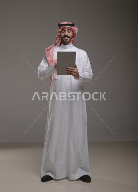 ترويج المنتجات التقنية الحديثة , بورتريه لرجل عربي خليجي سعودي يرتدي الزي  التقليدي يحمل بيده  التابليت   , الضغط والنقر باستخدام حاسة اللمس , التسويق الرقمي و الإعلانات  ، تطبيقات الأجهزة اللوحية  ، صورة الجسم بالطول الكامل , خلفية رمادية .