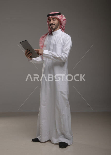 مفهوم الإعلانات والتسويق الالكتروني , بورتريه لرجل عربي خليجي سعودي يرتدي الزي  التقليدي يحمل بيده  التابليت و يشير بأصبع السبابة اتجاه الجهاز ،عرض خدمات الأجهزة التقنية الحديثة , الضغط والنقر باستخدام حاسة اللمس  ، برامج وتطبيقات الأجهزة اللوحية  ، صورة الجسم بالطول الكامل , خلفية رمادية .