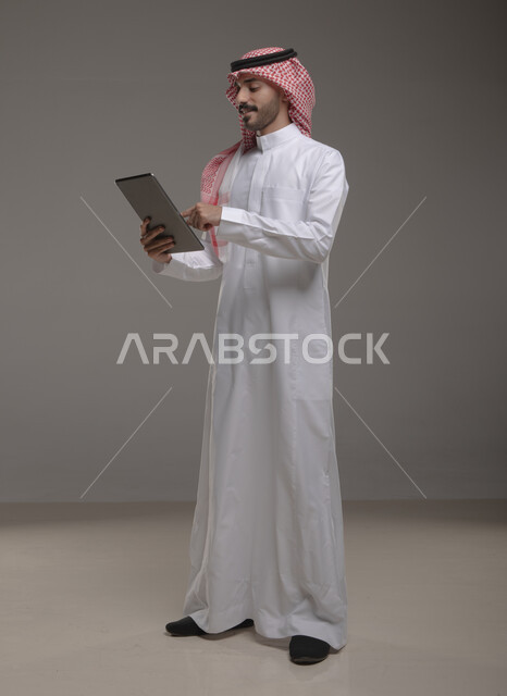 النقر والضغط باستخدام حاسة اللمس , بورتريه لرجل عربي خليجي سعودي يرتدي الزي  التقليدي يحمل بيده  التابليت  ،التسويق الرقمي و ترويجات إعلانية  ،برامج و  تطبيقات الأجهزة اللوحية  ، صورة الجسم بالطول الكامل , خلفية رمادية .